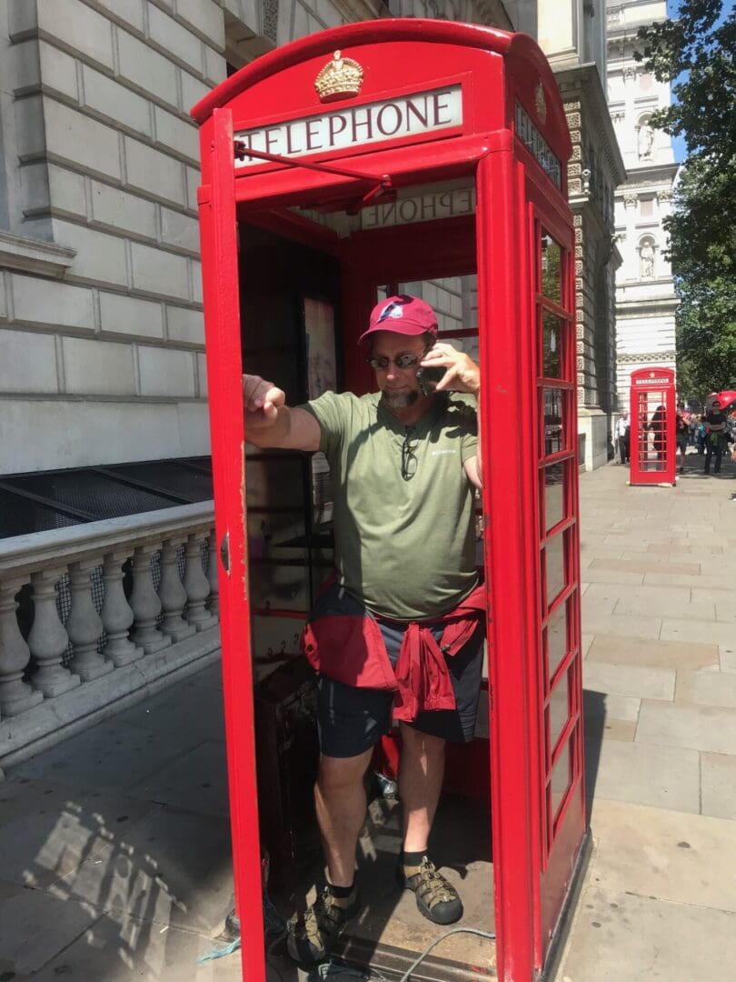 A man in a red phone booth on the sidewalk.